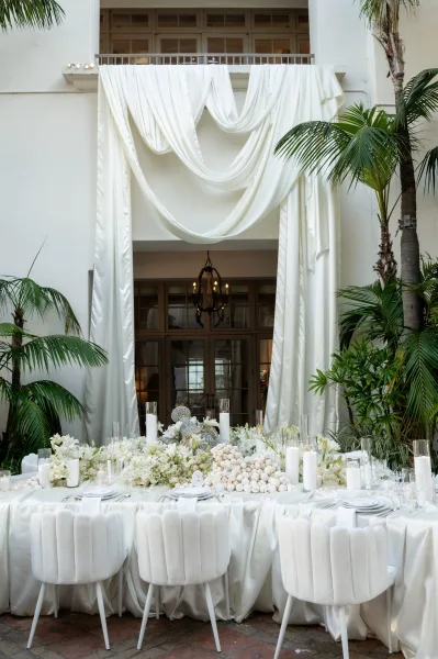 Reception tablescape with all-white reception decor, accented by white florals, below a chandelier amid palm plants and french doors.