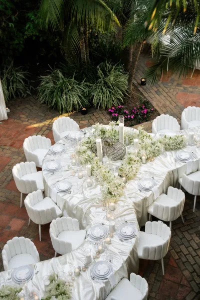 Reception tablescape on an outdoor reception table with a lush floral garland, set on a brick patio framed by tropical palms.