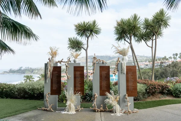 Seating chart display featuring a wedding seating chart, trimmed with pampas grass, set before an ocean view and palm trees.