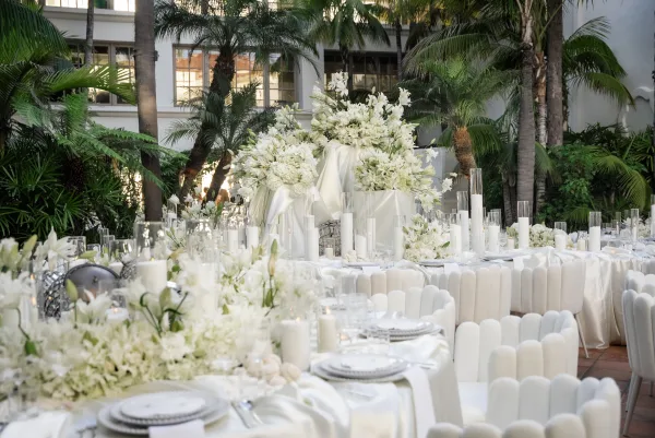 Reception tablescape with white floral centerpieces and satin table linens arranged in a tropical courtyard framed by palm trees.