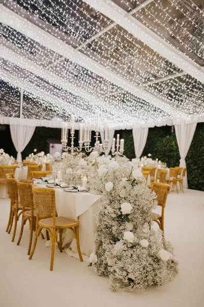 White roses and baby's breath decorate a reception tablescape, with black ribbon napkins, candelabras and rattan chairs beneath string lights