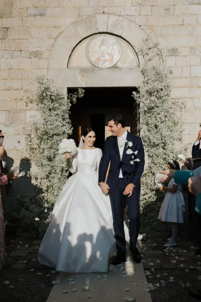 Newlyweds walk hand in hand through a wooden church doorway as rose petals fall around them**