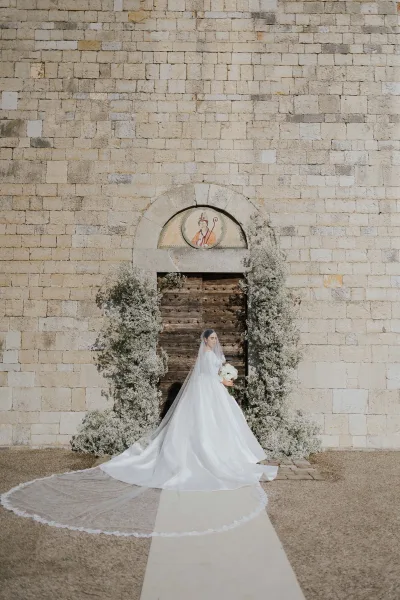 Structured satin gown, lace-trimmed cathedral veil and white roses appear in a bridal portrait beneath a baby's-breath arch