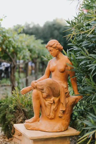 Terracotta garden statue depicts a seated woman amid dense greenery beside a pale gravel path