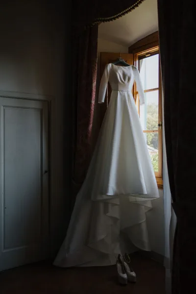 White satin wedding dress hangs from a hanger beside a curtained window, with its long sleeves clearly visible