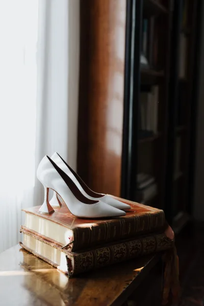 White pointed-toe bridal pumps with sculptural heels rest on vintage books atop a wooden table beside a sheer curtain