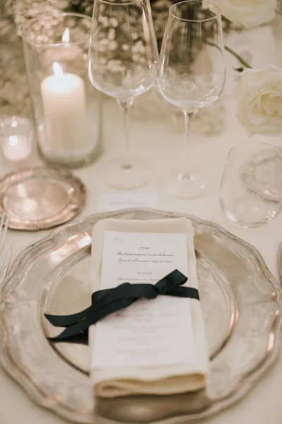 Silver charger plate, ribbon-tied menu card and folded linen napkin form a wedding place setting beside white roses