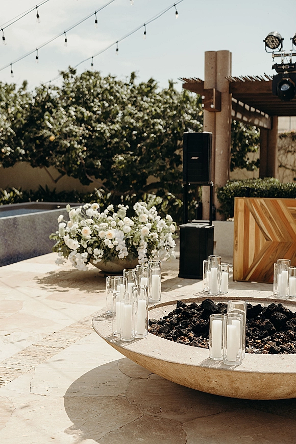 Reception lounge decor with a fire pit, glass cylinder candles, and white floral arrangement under string lights on a stone patio pergola