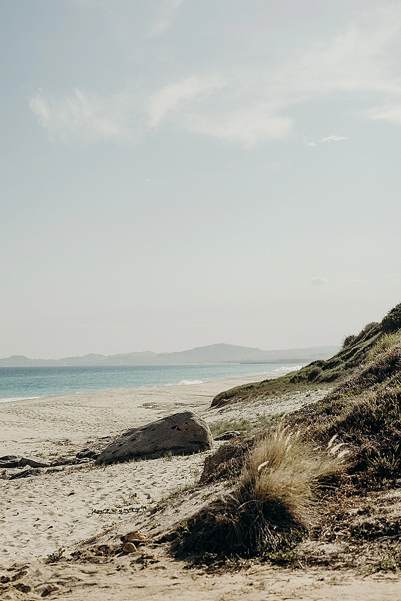 Beach landscape with empty beach shoreline, ocean horizon beyond sandy dunes and coastal grass under soft clouds and distant hills