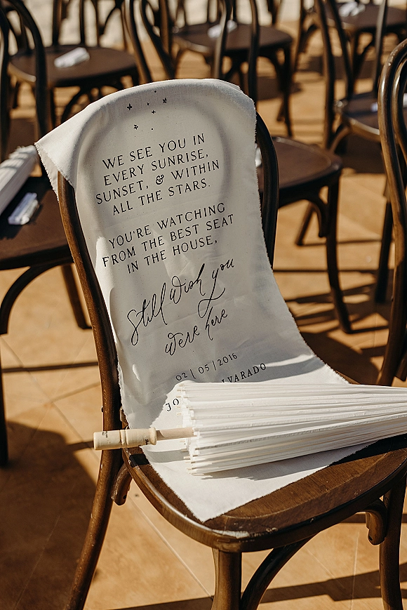 Wedding memorial chair with an in loving memory chair sign and fabric drape, set among wooden ceremony seating rows on a wood floor