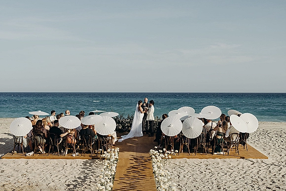 Wedding ceremony on beach with white parasols as bride and groom exchange vows beside a floral wooden aisle runner, ocean horizon behind