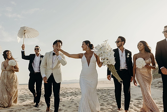 Wedding party portrait of the bride and groom with beach wedding party walking on sand, bride holding a white orchid bouquet by the ocean horizon