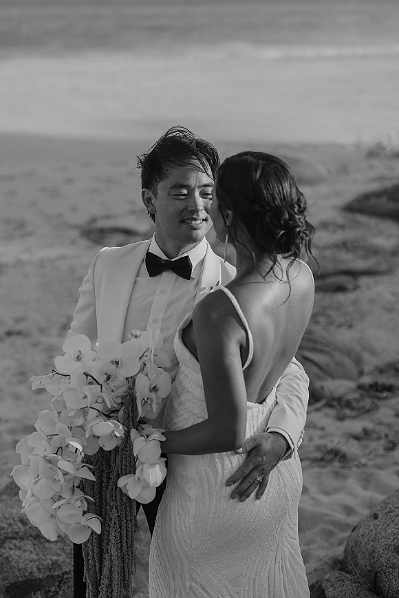 Couple portrait in a black and white wedding portrait, bride and groom embracing on a rocky beach with ocean backdrop and orchid bouquet