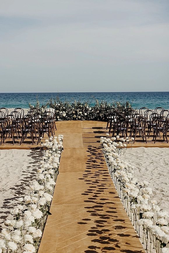 Ceremony aisle design with a runner on sand and rows of white flowers beside wooden chairs, set against the ocean horizon sky