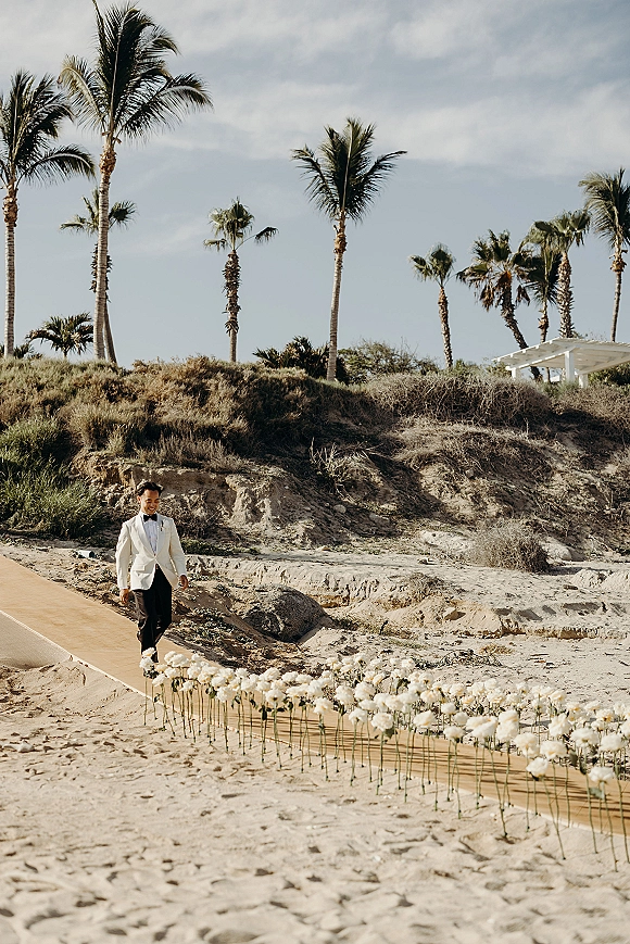 Groom portrait of a groom walking down aisle in a white tuxedo jacket and black bow tie on a wooden beach runner with white flowers, dunes and palms behind