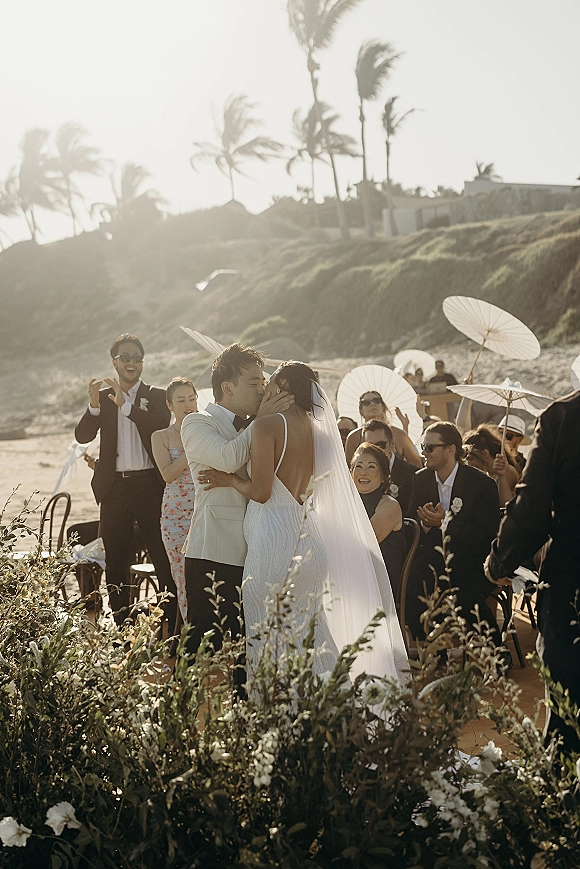Wedding kiss at a beach wedding ceremony as bride and groom embrace, veil blowing, guests with white parasols by ocean cliffs