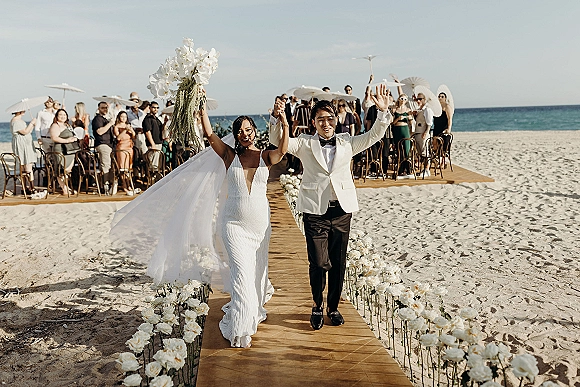 Wedding recessional as bride and groom walk down aisle holding hands, bouquet raised, by a sandy beach with ocean and guests cheering