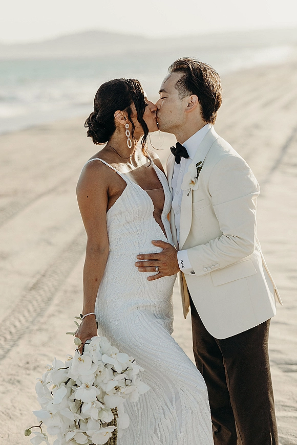 Wedding kiss portrait of bride and groom kissing on a sandy beach, bride holding an orchid bouquet with ocean shoreline and distant hills behind