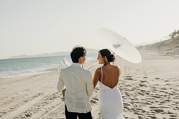 Couple portrait of bride and groom at a beach wedding couple, holding hands on sandy shoreline with ocean backdrop and white parasol