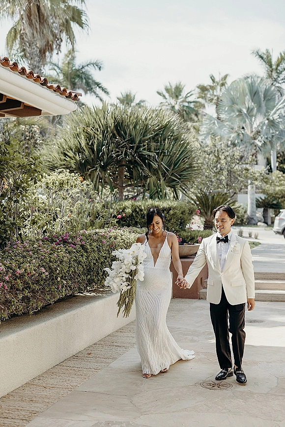 Couple portrait of bride and groom walking hand in hand, her cascading orchid bouquet against palm-lined garden walkway by stucco steps