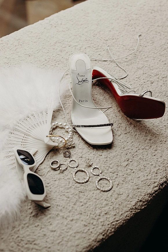 Bridal accessories flat lay with white strappy heels, pearl bracelet, diamond hoops, and wedding rings on textured fabric surface