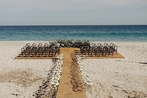 Beach wedding ceremony setup with oceanfront ceremony chairs flanking an aisle runner and white rose markers, facing a floral altar by the sea