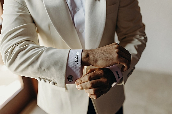 Groom getting ready adjusts groom cufflinks on a white French cuff shirt with a cream suit jacket, lit by soft window light against a neutral wall