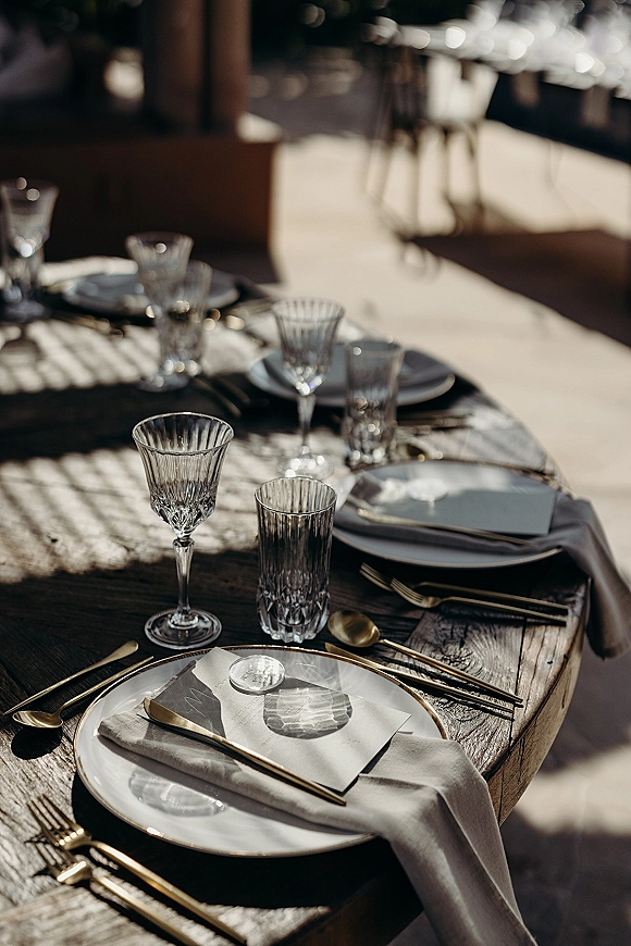 Reception tablescape with gold flatware, white plates, linen napkins, crystal goblets and wax-sealed place cards on a sunlit outdoor patio table