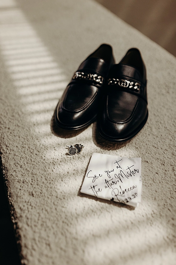 Groom accessories flat lay with black leather loafers, silver cufflinks, and a handwritten note on textured fabric in window light shadows