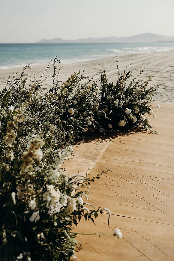 Ceremony aisle florals with beach wedding ceremony setup featuring white flowers and greenery garland on a wooden platform by the ocean