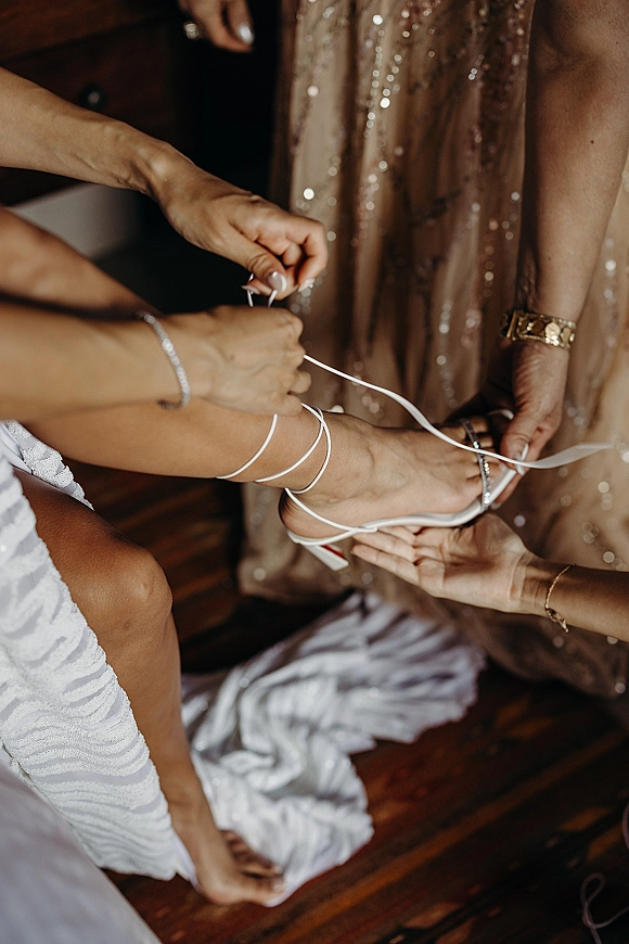 Bridal shoes as a helper ties the wedding shoe details, securing ribbon ankle wraps on strappy heels over a wood floor indoors