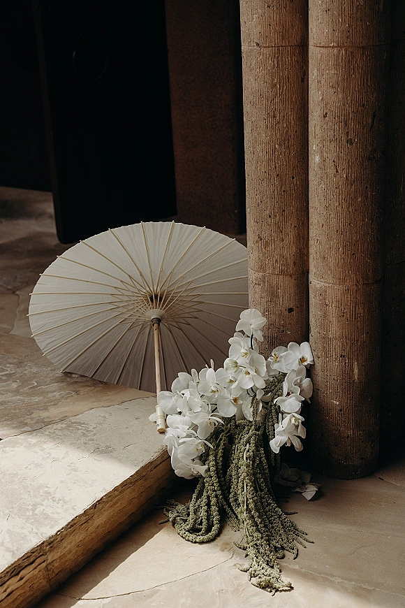 Wedding bouquet of white orchids with cascading hanging greenery on a stone floor beside a white paper parasol near a shadowed doorway