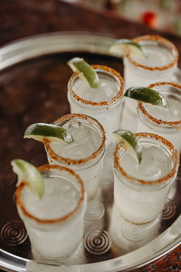 Margarita shots on a metal tray with tequila shot glasses, ice, lime wedges, and chili-salt rims on a bar surface