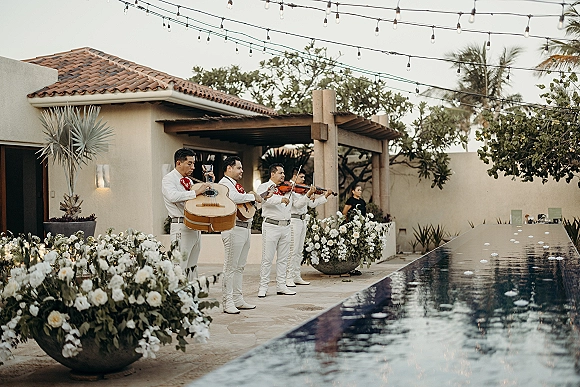 Wedding mariachi band performing with guitars and violin in suits by a pool under string lights in a stucco courtyard with palm trees