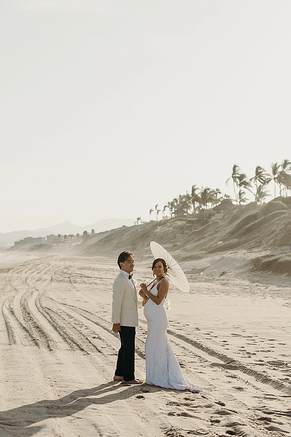 Couple portrait on a sandy beach, bride in a strapless wedding dress holding a parasol beside groom in a white dinner jacket, dunes and palms behind