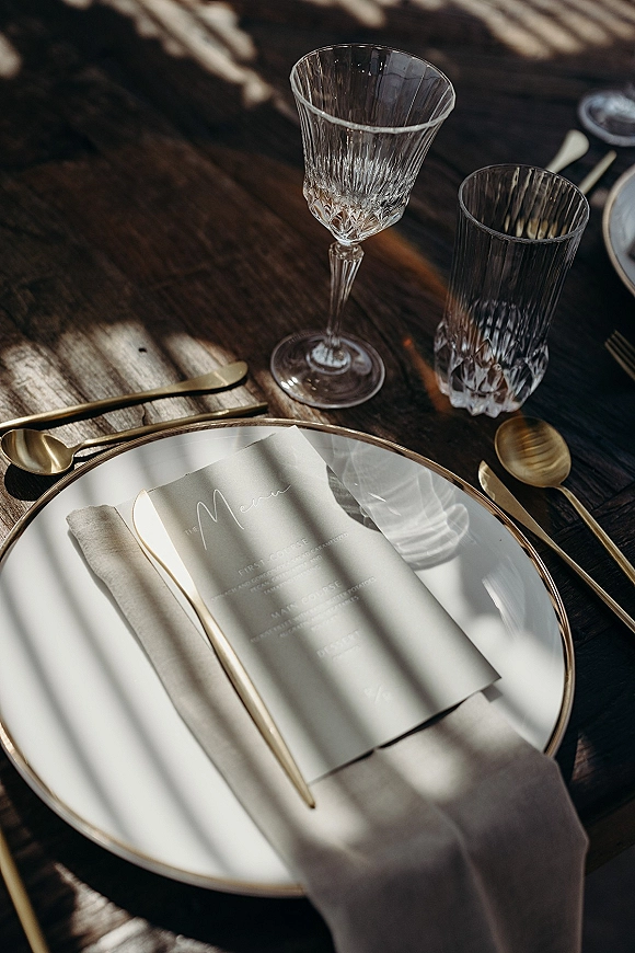 Wedding place setting with a wedding menu card on a dinner plate, beige napkin, gold flatware, and glassware on a dark wood table in dappled sunlight