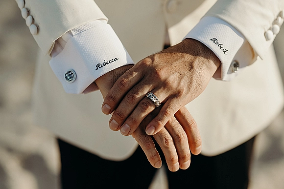 Wedding ring close-up of a hand wearing a diamond wedding band, with cufflinks and embroidered shirt cuffs against a neutral backdrop