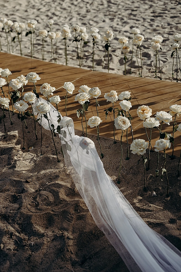 Ceremony aisle decor on a beach wedding aisle, white roses on stakes and sheer fabric draping leading to a wooden platform in sand