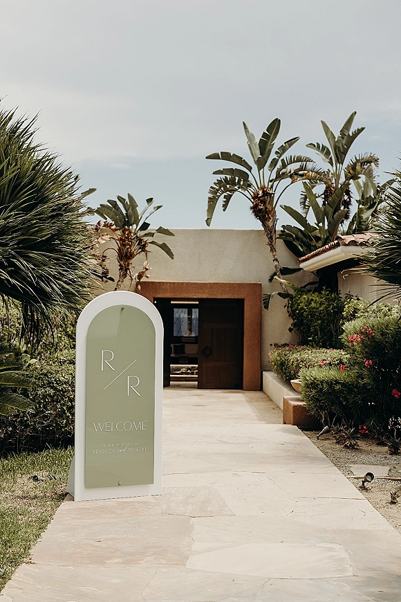 Wedding welcome sign on an acrylic stand with monogram lettering along a stone walkway by a stucco doorway, palms and shrubs behind