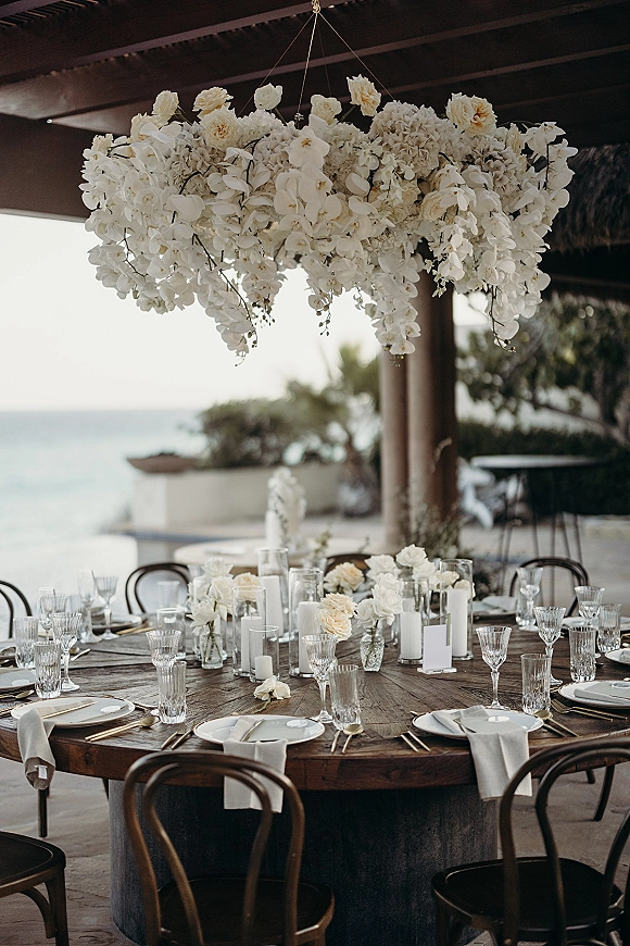 Reception tablescape with hanging floral centerpiece of white orchids, hydrangeas, and roses above a candlelit round wooden table on an ocean-view terrace