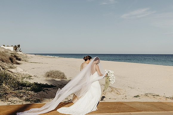 Bridal portrait of a bride with long veil holding a bouquet on a beach, her cathedral veil trailing over sand dunes by the ocean