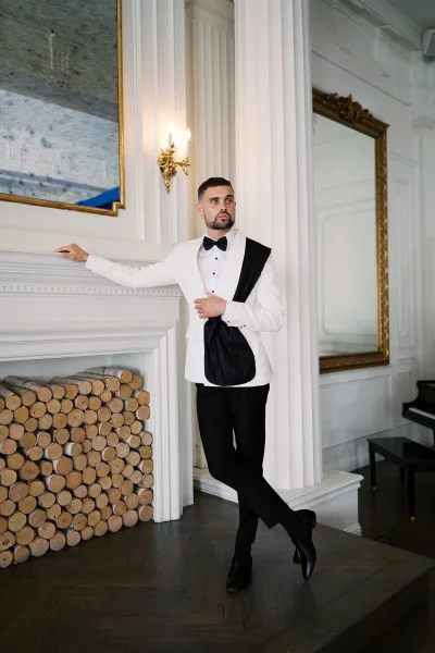 Groom portrait in a white dinner jacket groom look, wearing a black bow tie and boutonniere, posed by a fireplace and mirror in a paneled room