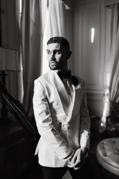 Groom portrait in a white tuxedo jacket with black bow tie and boutonniere, posed by window light in an elegant indoor room