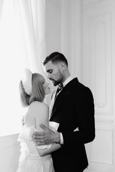 Wedding couple portrait in a close embrace, bride in a strapless dress with headpiece and groom in a black tuxedo by a white paneled wall
