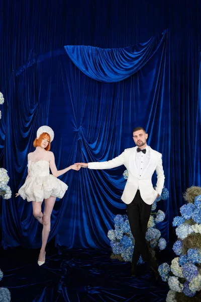 Couple portrait of bride and groom holding hands, her short wedding dress and headpiece against a blue draped studio backdrop with hydrangeas