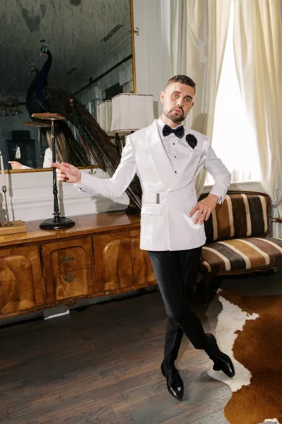 Groom portrait in a white tuxedo jacket with black bow tie, standing by a mirror and striped sofa in a vintage room with window curtains