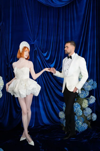 Couple portrait of bride and groom holding hands, bride in short wedding dress with headpiece before blue velvet curtains and hydrangeas