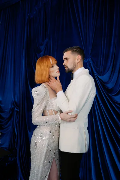Couple portrait of bride and groom face to face, groom holding her cheeks; bride in sequin gown with earrings before blue draped backdrop