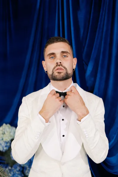 Groom portrait in a white tuxedo jacket adjusting a black bow tie, wearing a boutonniere against blue draped fabric and florals