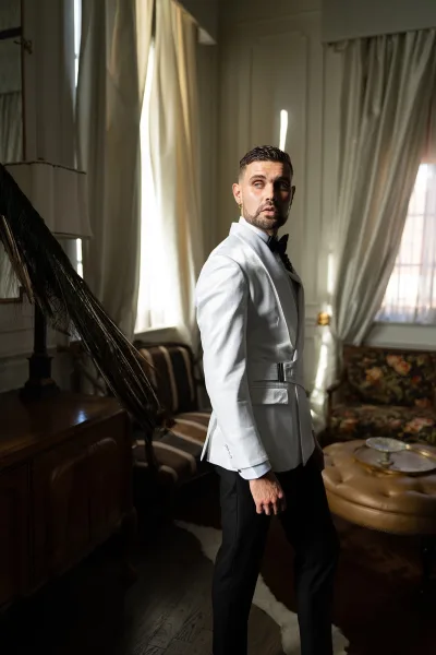 Groom portrait in a white tuxedo jacket with black bow tie and boutonniere, standing by curtained windows in a vintage lounge